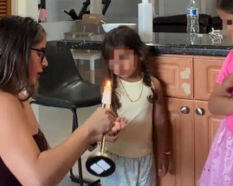 Woman holding a lit candle toward a small girl in a kitchen, while another child watches nearby.