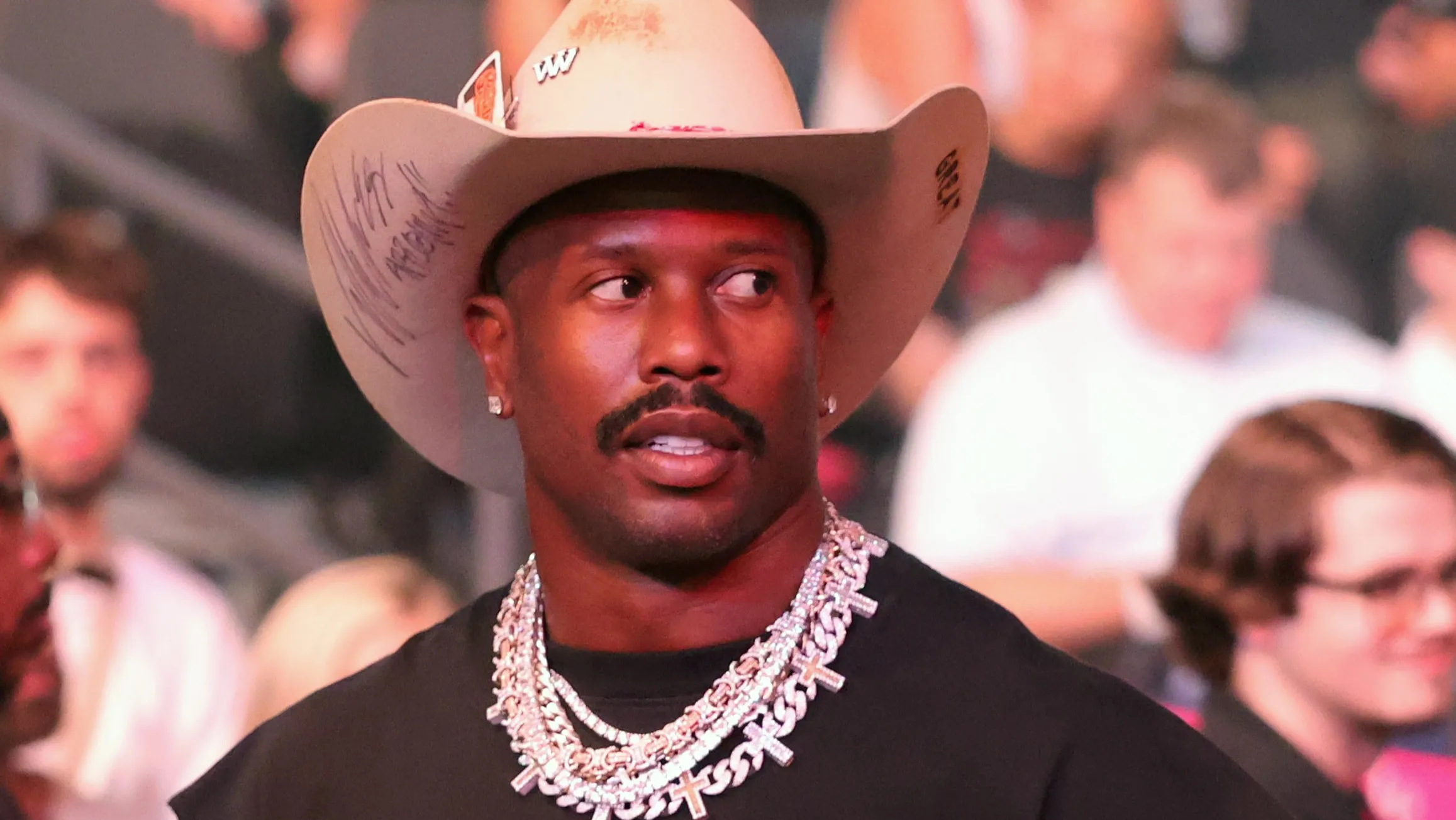 Man wearing a beige cowboy hat and heavy silver jewelry, looking to the side at a crowded event.
