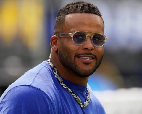 Man wearing sunglasses and a silver chain necklace, smiling outdoors in a blue shirt.