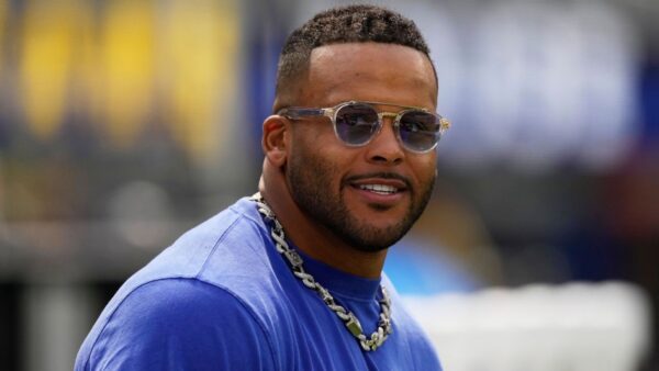 Man wearing sunglasses and a silver chain necklace, smiling outdoors in a blue shirt.