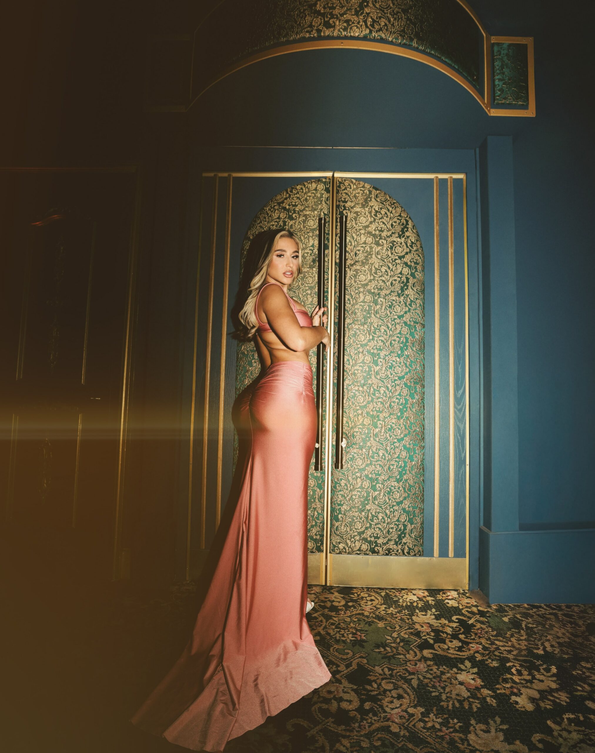 Glamorous woman in a pink satin gown poses beside an ornate teal and gold door in a dimly lit room.