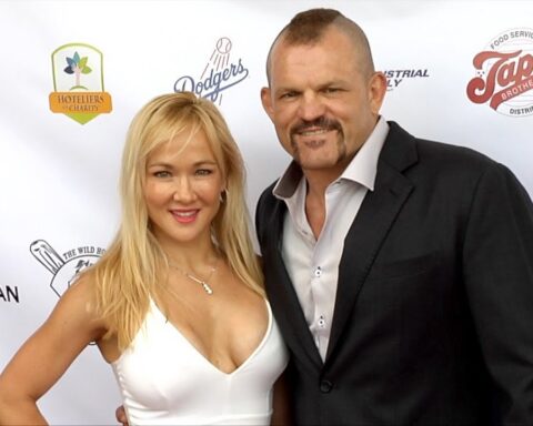 Two adults smiling for a photo on a red carpet, woman in a white dress and man in a dark suit with sponsor logos behind them.