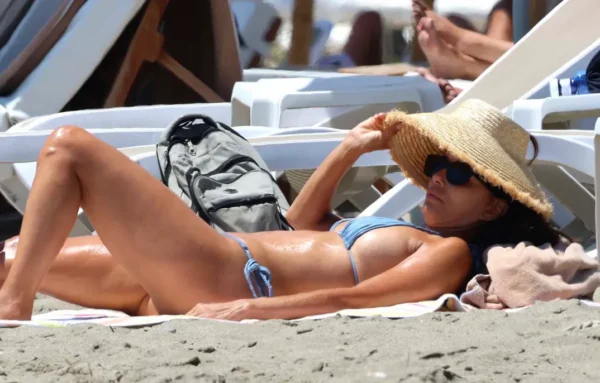 Woman in a blue bikini sunbathing on the beach under a straw hat and sunglasses, resting on a towel with a backpack nearby
