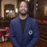 Man in a blue blazer standing in a warmly lit bar, facing the camera with a small badge on the blazer pocket