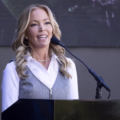 Woman speaking at a podium with a microphone on stage, wearing a white shirt and gray vest, smiling.