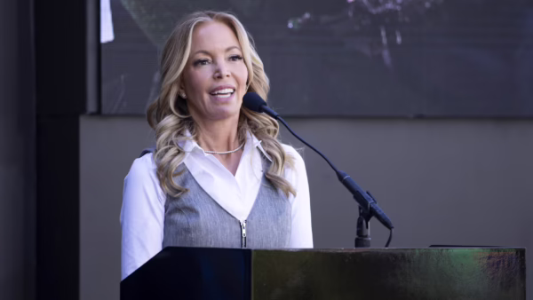 Woman speaking at a podium with a microphone on stage, wearing a white shirt and gray vest, smiling.