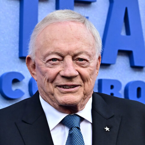 Older man in a dark suit and blue patterned tie standing before a blue backdrop with large lettering behind him.