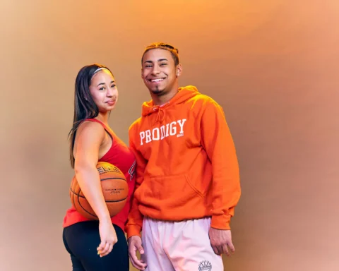 Two young adults pose together in athletic wear, the woman holding a basketball, against a warm orange backdrop.