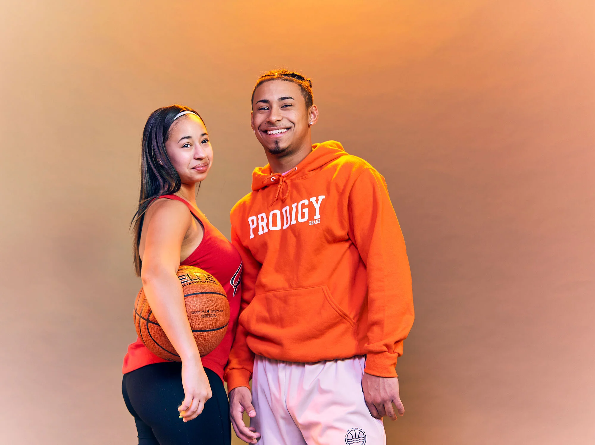 Two young adults pose together in athletic wear, the woman holding a basketball, against a warm orange backdrop.