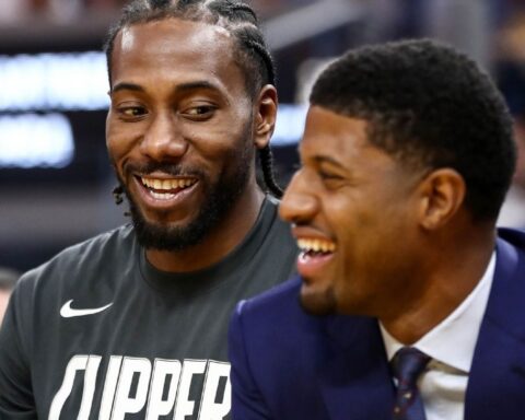 Two men smiling and chatting at a basketball game; one in a Clippers shirt, the other in a dark suit and tie, both laughing.