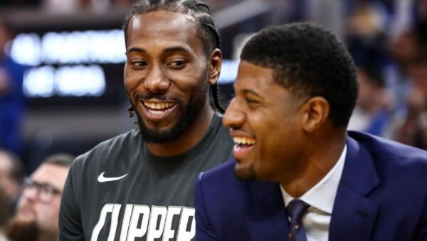 Two men smiling and chatting at a basketball game; one in a Clippers shirt, the other in a dark suit and tie, both laughing.