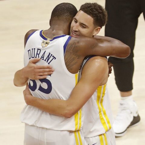 Two basketball players in white and gold jerseys hug on the court, showing team camaraderie after a game (one jersey reads 'DURANT').