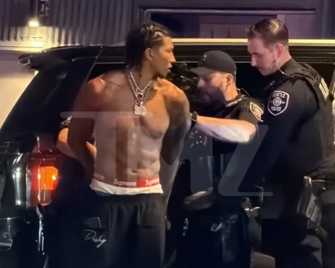 Shirtless muscular man with braided hair and chain jewelry talks to two uniformed officers beside a black SUV at night.
