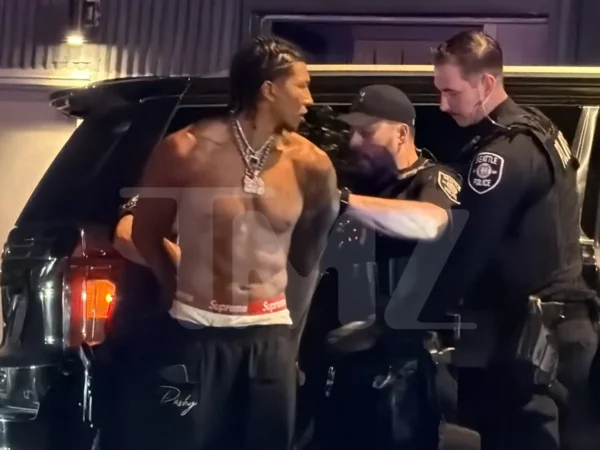 Shirtless muscular man with braided hair and chain jewelry talks to two uniformed officers beside a black SUV at night.