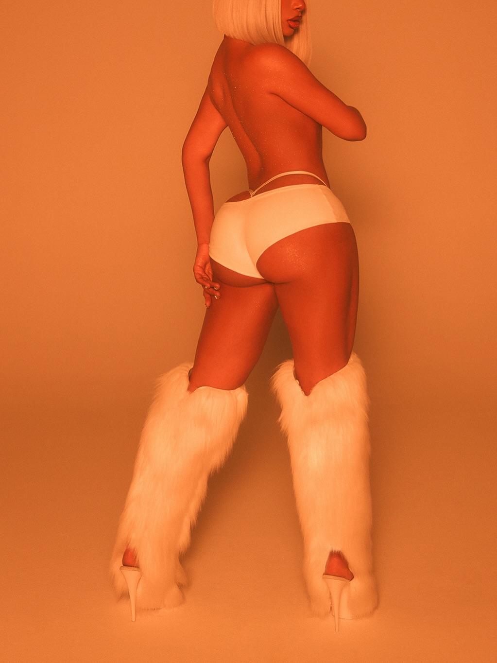 Back view of a topless person wearing white underwear and fluffy white fur leg warmers with high heels; orange studio lighting.