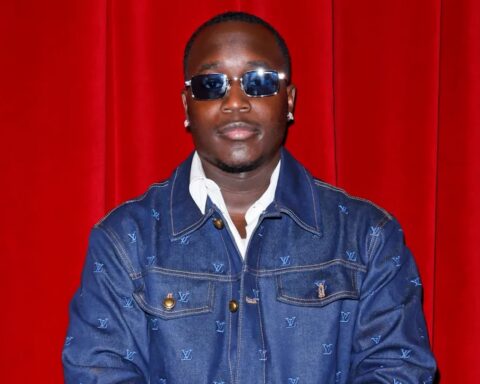 Portrait of a man wearing sunglasses and a denim LV jacket in front of a red curtain.