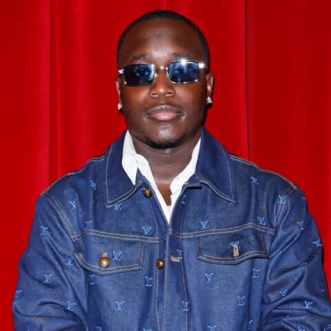 Portrait of a man wearing sunglasses and a denim LV jacket in front of a red curtain.