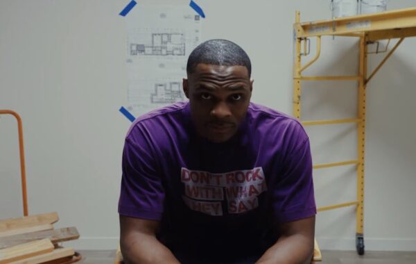 Man in a purple T‑shirt sits in a workshop, facing the camera, with blue taped architectural plans on the wall and a yellow scaffold to the right.