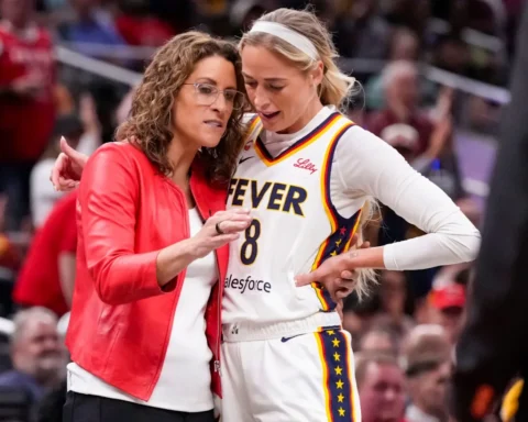 Basketball player in a white jersey with 'FEVER' talks with a woman in a red jacket on the sideline.