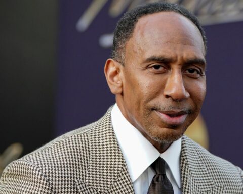Close-up portrait of a man in a brown houndstooth blazer and white shirt at a formal event on a dark purple background.