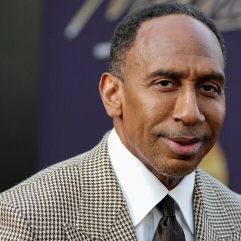 Close-up portrait of a man in a brown houndstooth blazer and white shirt at a formal event on a dark purple background.