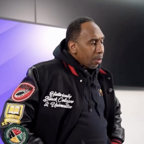 Man in a black varsity jacket with patches, looking down in a studio setting with a purple-lit background.