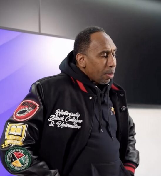 Man in a black varsity jacket with patches, looking down in a studio setting with a purple-lit background.