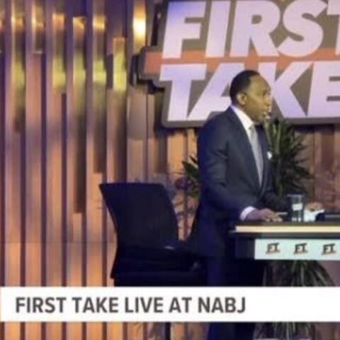 Man in a suit stands at a desk on the First Take set, NABJ live broadcast backdrop behind him.