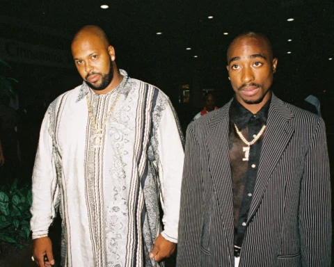 Two men in stylish outfits pose together at a nighttime event; one wears a light patterned robe-like shirt, the other a pinstripe suit with gold chains visible.