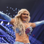 Wrestler in a glittery blue outfit raises both arms in the ring as stage lights glow and confetti-like lights swirl around her.