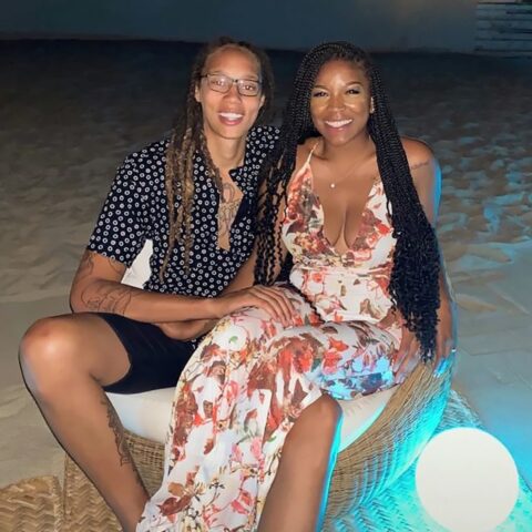 Two people sit closely on wicker chairs at the beach at night, smiling for the camera; one wears a black patterned shirt and shorts, the other a floral dress with braided hair.