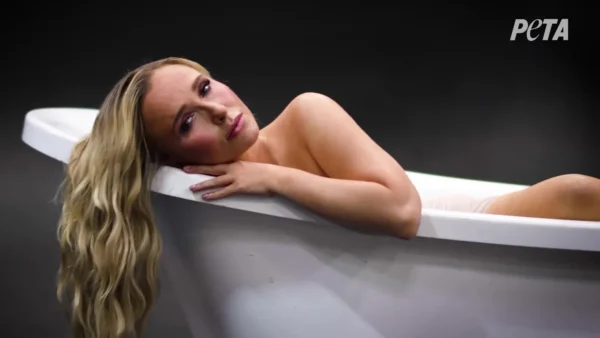 Nude woman lying in a white bathtub, head resting on the edge, gazing toward the camera; PETA logo appears in the top-right corner.