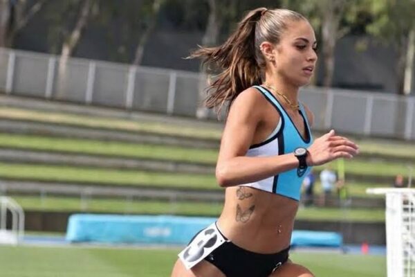 Female runner sprinting on a track in a blue and black sports top and black shorts, bib number 8 visible, ponytail flying and a hip tattoo showing.