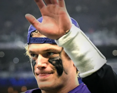 Athlete in a purple jersey with a backwards cap raises a hand in celebration, with black eye paint visible under the eyes.