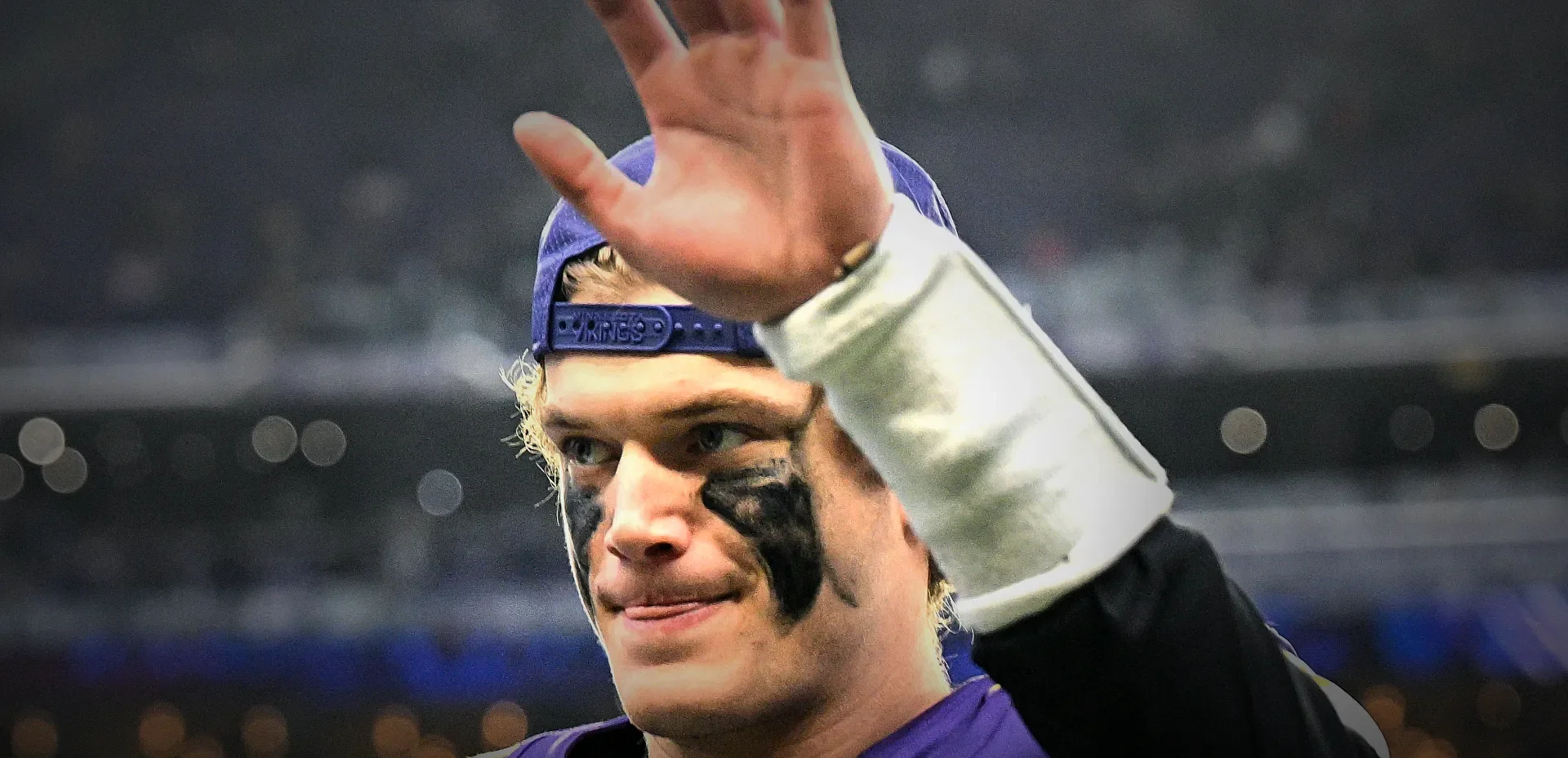 Athlete in a purple jersey with a backwards cap raises a hand in celebration, with black eye paint visible under the eyes.